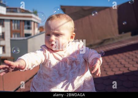Primo piano di cute baby girl indossare adorabile vestito sul tetto, bolle di sapone galleggianti intorno, bambina cercando di prendere le bolle Foto Stock