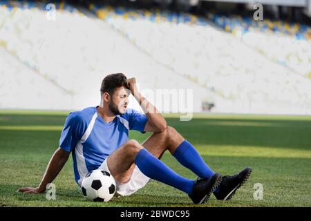 triste giocatore professionista di calcio in uniforme blu e bianca seduto con palla sul campo di calcio allo stadio Foto Stock