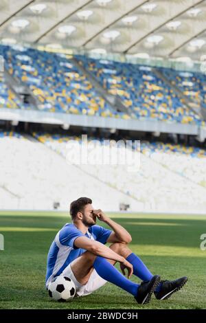 triste giocatore professionista di calcio in uniforme blu e bianca seduto con palla sul campo di calcio allo stadio Foto Stock