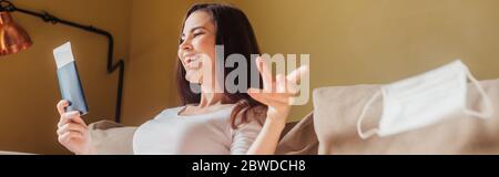 raccolto panoramico di donna allegra gettando in aria maschera medica e tenendo passaporto con biglietto aereo, fine del concetto di quarantena Foto Stock