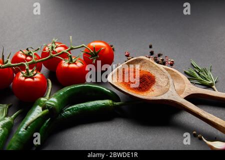 pomodori ciliegini rossi, rosmarino, pepe, cucchiai di legno con paprika in polvere e peperoncini verdi su nero Foto Stock