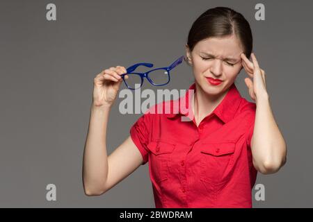 Bella ragazza giovane in una camicia rossa che ha occhi irritati e una testa da occhiali su uno sfondo grigio Foto Stock