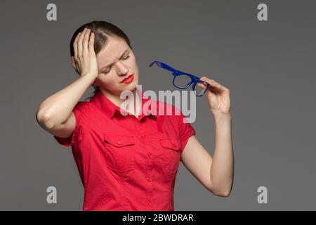 Bella ragazza giovane in una camicia rossa che ha occhi irritati e una testa da occhiali su uno sfondo grigio Foto Stock