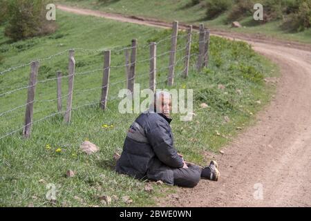 Una stanca rural uomo seduto accanto alla strada sul pavimento, una fattoria è racchiusa da un recinto, Iran, Gilan Foto Stock