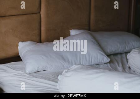 Un letto disordinato con due cuscini e due cuscini per il corpo e lenzuola di cotone, tutti in bianco, con uno sfondo beige dietro il letto. Foto Stock