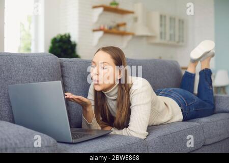 Datazione online. Donna che manda bacio d'aria e usa il computer portatile in salotto chat room. Foto Stock