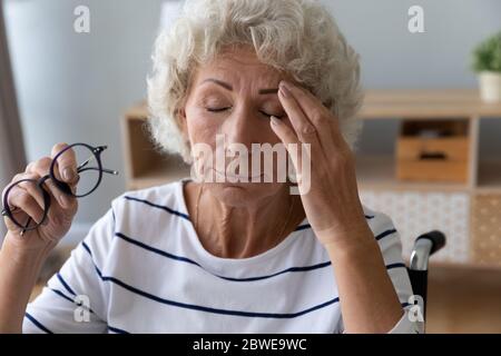 Donna anziana seduta in sedia a rotelle togliendo gli occhiali riduce la pioggia oculare Foto Stock