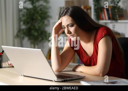 Donna preoccupata che legge sul computer portatile seduto su una scrivania di notte a casa Foto Stock