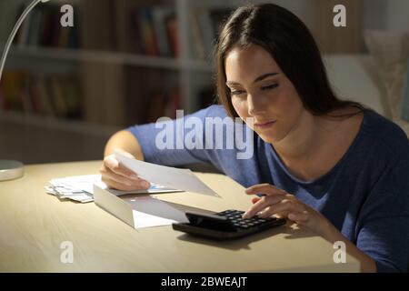 Donna seria che calcola le ricevute sulla calcolatrice seduta su un tavolo di notte a casa Foto Stock