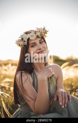 Bella giovane donna brunnete in corona e khaki vestire camminare in campo e raccogliere fiori. Tramonto estivo. Stile di vita in campagna. Stile rustico. Foto Stock