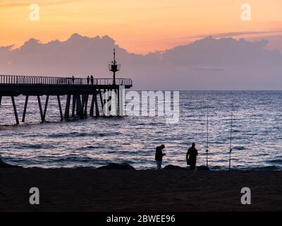 Due pescatori pescano sulla spiaggia al tramonto utilizzando canne da pesca, adagiato contro la costa. Foto Stock
