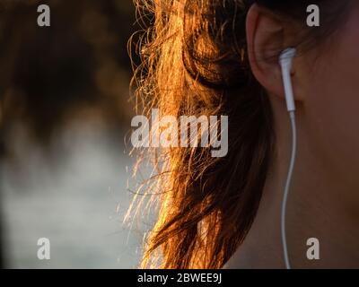 Una ragazza con una bella luce solare in capelli sta ascoltando musica con gli auricolari. Nessuna faccia. Foto Stock