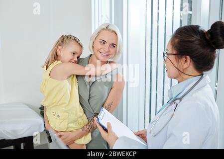 Bella giovane donna sorridente che porta la sua figlia piccola quando si parla con pediatra su diagnosi e prescrizione Foto Stock