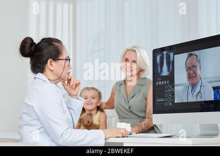 Pediatra pensiva serio avendo consultazione in linea con pulmonologa e discutendo i polmoni x-ray di bambina Foto Stock