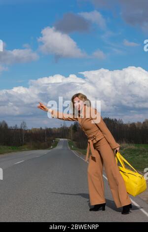 Elegante donna di mezza età in un abito alla moda e con una borsa da viaggio gialla in mano attraversa la strada. Il concetto di riposo dalla vita cittadina e. Foto Stock