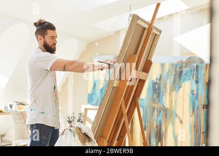 Vista laterale ritratto di artista maschile contemporaneo pittura immagine su cavalletto mentre si lavora in studio d'arte illuminato al sole, copia spazio Foto Stock