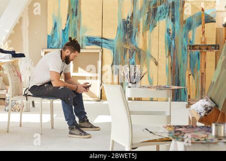 Vista laterale completa dell'artista con bearded contemporaneo che usa lo smartphone mentre si siede sulla sedia in studio d'arte con dipinti astratti sullo sfondo, copia Foto Stock