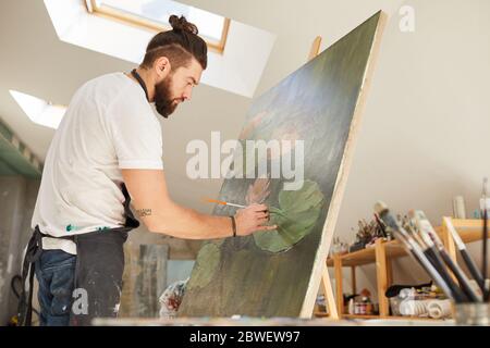 Vista laterale ritratto di talento artista maschile pittura immagine sul cavalletto mentre lavora in uno spazioso studio d'arte illuminato dalla luce del sole, copia spazio Foto Stock