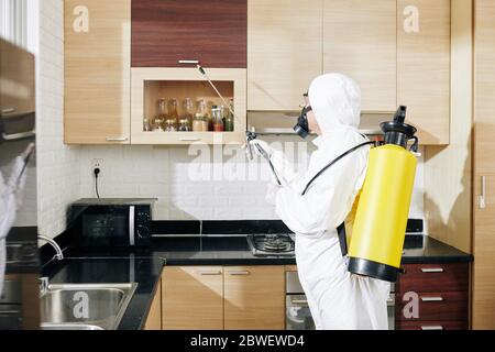 Disinfettante anti-coronavirus operaio in tuta di nocciolo applicando spray su tutte le superfici in cucina del cliente Foto Stock