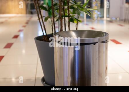 primo piano contenitore in acciaio cilindro contenitore rifiuti. vicino in sfocatura una pianta in una vasca grigia. Sullo sfondo di una piastrella pavimento chiaro e l'interno di un pubblico Foto Stock