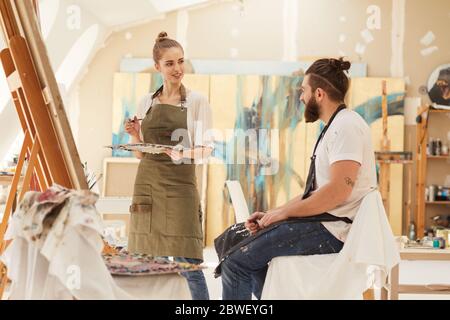 Ritratto di giovane donna pittura immagine sul cavalletto mentre si lavora in studio d'arte illuminato al sole con uomo bearded seduto su sedia e posa, copia spazio Foto Stock