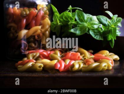 Mazzo di foglie di basilico fresco verde posto vicino al vaso e un mucchio di pasta colorata cruda sul tavolo Foto Stock
