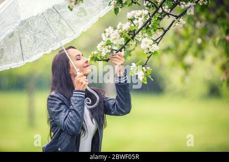 La donna odori fiori di albero su durante una giornata di pioggia primaverile nella natura, tenendo un ombrello. Foto Stock