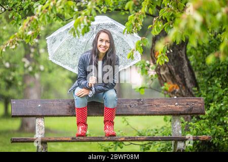 Sorridente bella donna che tiene ombrello sulle spalle si siede su una panchina-riposo nel parco sotto la pioggia. Foto Stock