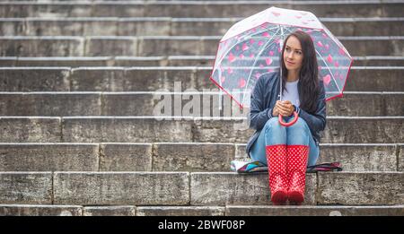 La ragazza si siede aspettando sulle scale di pietra, indossando stivali da pioggia rossi e un ombrello con i cuori che la proteggono contro la pioggia. Foto Stock