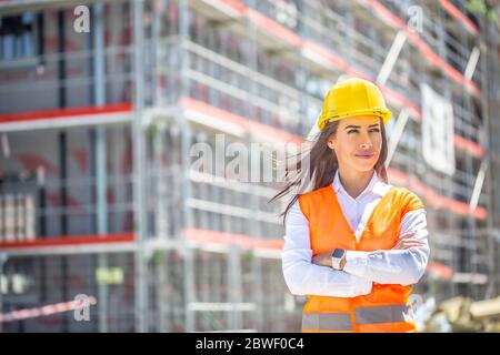 Supervisore del sito femminile che indossa gilet di sicurezza e casco si trova in posizione sicura di fronte alla costruzione coperta da ponteggi. Foto Stock