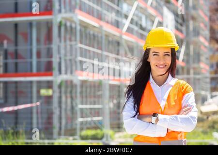 Supervisore del sito femminile che indossa gilet di sicurezza e casco si trova in posizione sicura di fronte alla costruzione coperta da ponteggi. Foto Stock