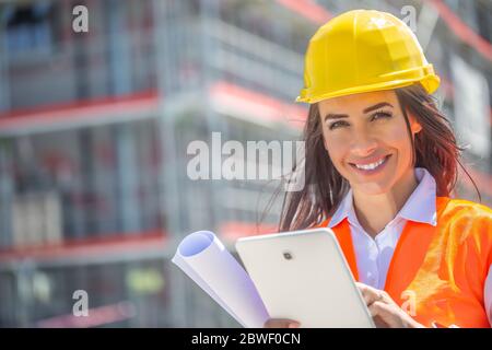 Donna sorridente in PPE tiene un tablet e un progetto con un cantiere in background. Foto Stock