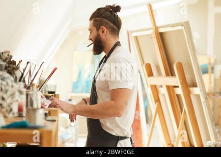 Ritratto di un artista bearded contemporaneo che sceglie strumenti e pennelli mentre lavora uno studio d'arte illuminato dalla luce del sole, spazio di copia Foto Stock