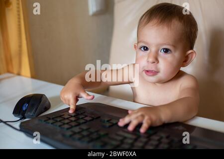 piccolo bambino carino seduto su una sedia da ufficio, guarda nella fotocamera e clicca su entra pulsante sulla tastiera, primo piano, messa a fuoco morbida Foto Stock