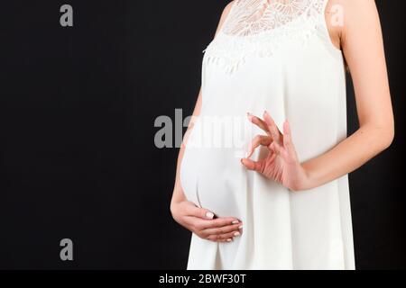 Immagine tagliata di donna incinta positiva in abito bianco che mostra un gesto corretto contro il ventre su sfondo nero. Gravidanza facile e felice. Centro benessere copie Foto Stock