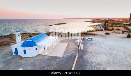 Vista aerea del tramonto sulla costa e tradizionale cappella dipinta di bianco con porte blu sulla spiaggia di Agia Thekla, Ayia Napa, Famagosta, Cipro da A. Foto Stock