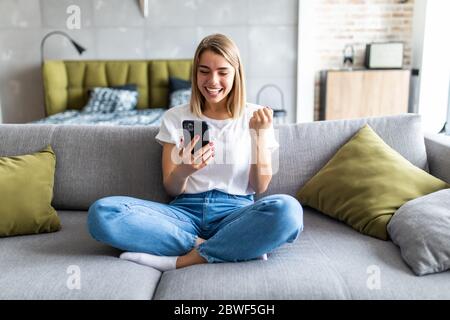 Donna entusiasta che guarda i contenuti multimediali online su un telefono cellulare seduto su un divano nel soggiorno Foto Stock