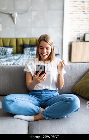 Donna entusiasta che guarda i contenuti multimediali online su un telefono cellulare seduto su un divano nel soggiorno Foto Stock