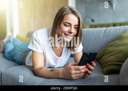 Piuttosto giovane donna sdraiata sul divano e scrivere messaggi con il suo smartphone Foto Stock