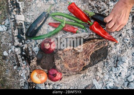 Verdure e carne alla griglia. Man Hand prepara la bistecca Ribeye con peperoni, cipolle e melanzane per cucinare sulla pietra all'aperto al fuoco Foto Stock