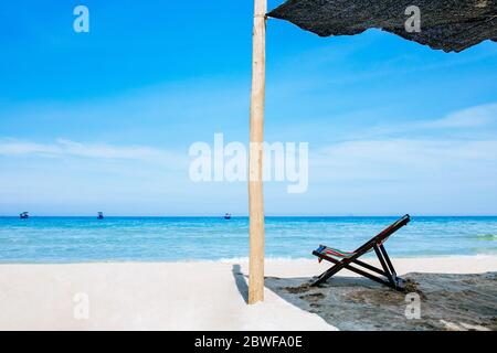 Una sedia a sdraio si trova all'ombra sulle rive di una spiaggia di sabbia sul mare. Il colore bruoso dell'acqua e del cielo blu con nuvole rotte nella b Foto Stock