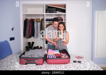Giovane coppia caucasica guardando lo smartphone sorridendo, preparando un viaggio. Passaporti e occhiali da sole sul letto. Concetto di imballaggio. Foto Stock