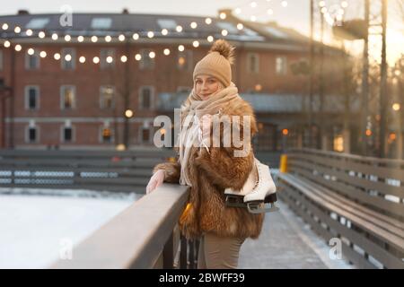 Giovane donna allegra in piedi e tenendo pattini di ghiaccio bianchi in giorno gelido, vista laterale, pista di ghiaccio su sfondo, all'aperto. Sport invernali Foto Stock