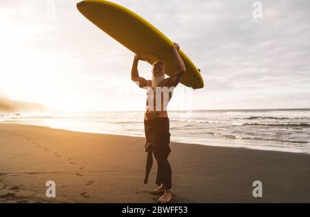 Tattooed surfer senior tenendo surf board sulla spiaggia al tramonto - ragazzo felice vecchio divertirsi facendo sport estremo - gioioso anziano concetto - Focus on hi Foto Stock