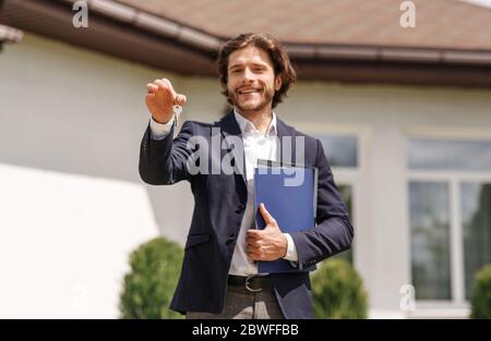 Sorridente agente immobiliare tenendo chiave casa vicino nuova residenza di vendita, all'aperto Foto Stock