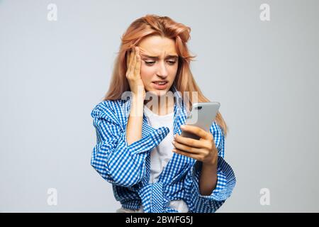 Scioccata sorpresa giovane cute donna con capelli biondi fragola guardando il suo telefono, riceve sms, notizie, messaggio, toccando la fronte, isolato su g. Foto Stock