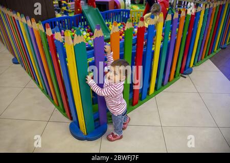 Un piccolo bambino carino si arrampica sulla colorata recinzione della sala giochi per bambini, guarda nella fotocamera e vuole giocare all'interno. Messa a fuoco morbida. Vista dall'alto, primo piano Foto Stock