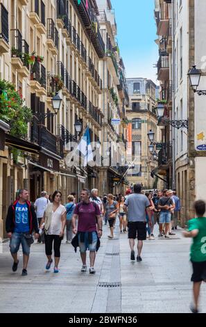 Turisti e gente del posto passeggiano lungo una delle strade cittadine di San Sebastian, in Spagna, in una giornata estiva luminosa con cielo blu chiaro. Foto Stock