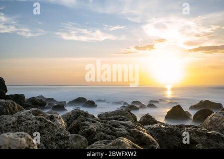 (Fuoco selettivo) splendida vista di una costa rocciosa bagnata da un mare liscio e setoso durante il tramonto. Melasti Beach con la sua scogliera in lontananza, Bali Sud. Foto Stock