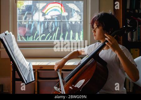 Un ragazzo focalizzato si siede di fronte alla finestra dipinta praticando violoncello Foto Stock